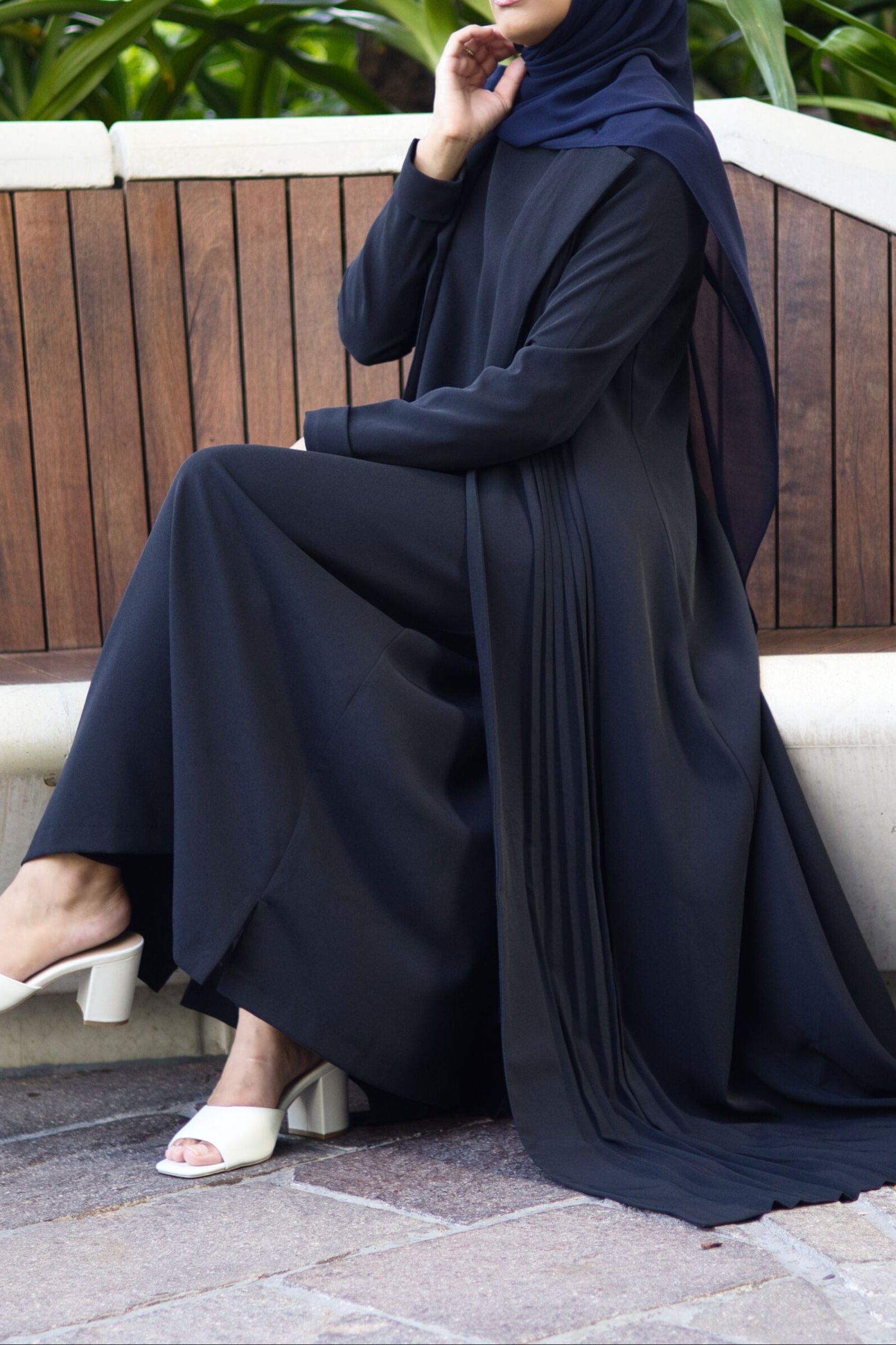 Muslim woman wearing the Amani Pleated Blazer Set by Modestly, seated outdoors on a wooden bench, styled in a navy hijab and white block heels. The modest, long-line blazer features pleated detailing, reflecting elegant and formal Islamic fashion.