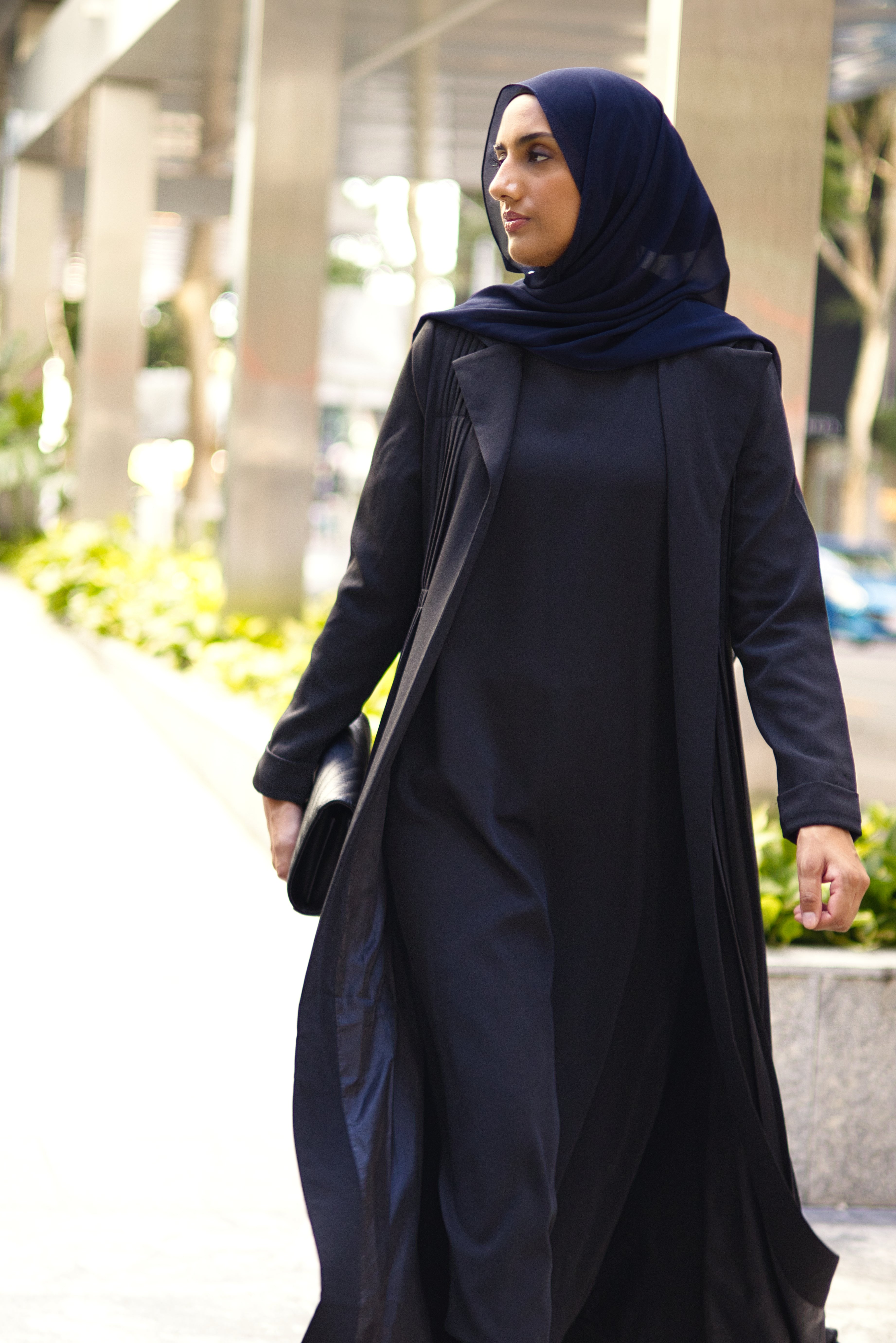 Hijabi woman confidently walking in black Amani Pleated Blazer Set, modest formalwear styled with clutch for a city look