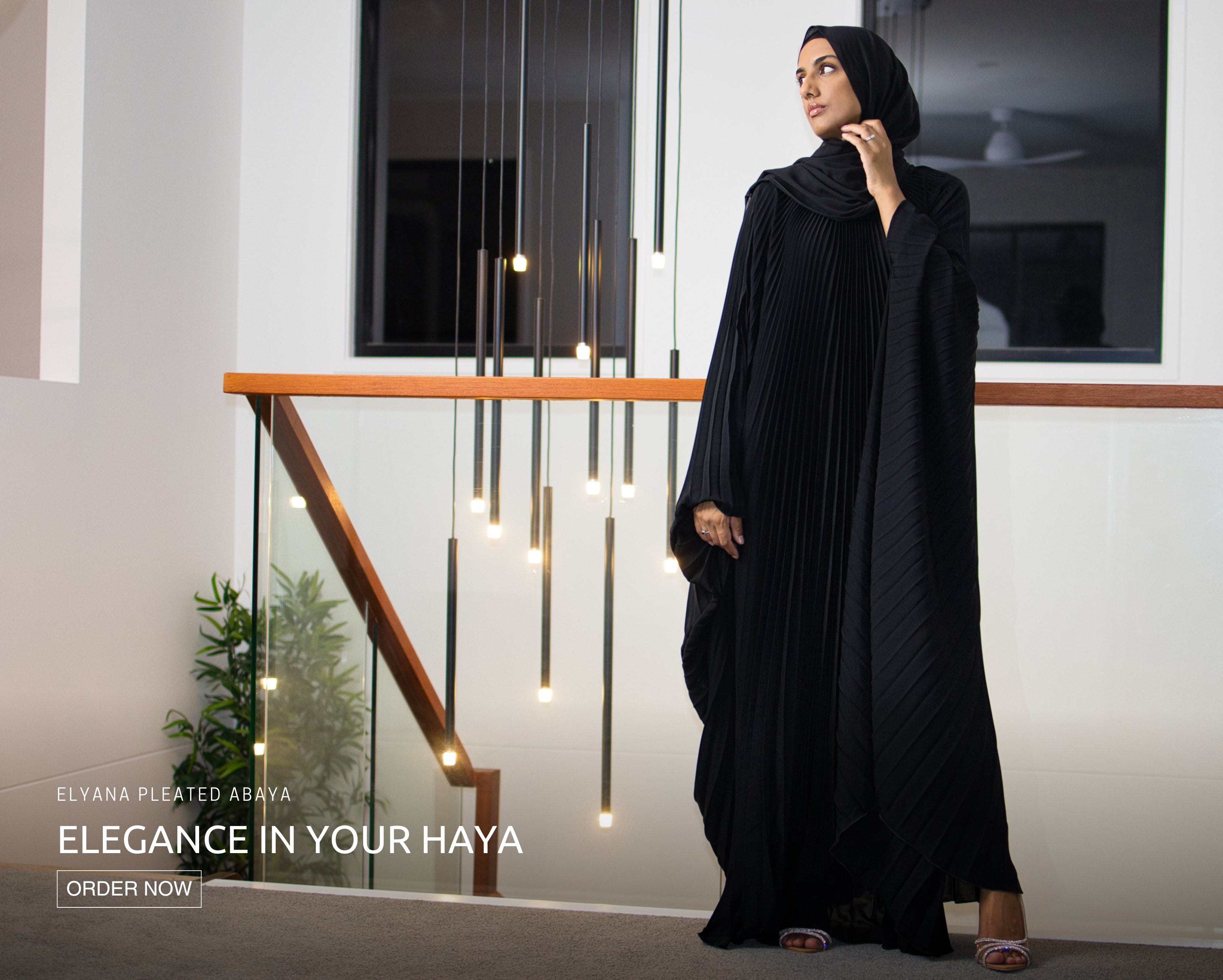 Muslim woman wearing the black Elyana Pleated Abaya, standing indoors under elegant hanging lights, showcasing graceful and modest Islamic evening wear.