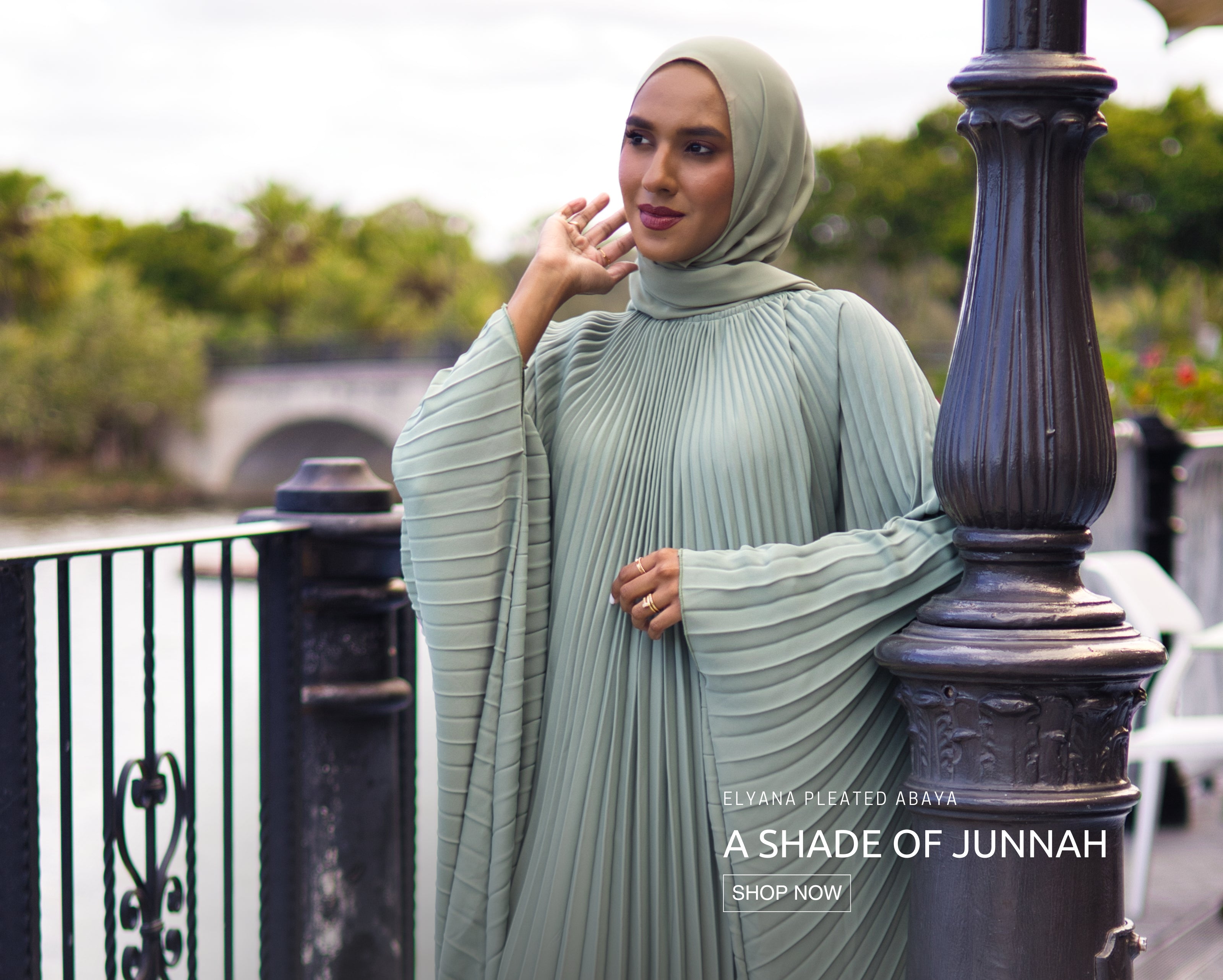 Muslim woman wearing sage green Elyana Pleated Abaya standing by a riverside railing, showcasing elegant modest fashion inspired by Jannah.